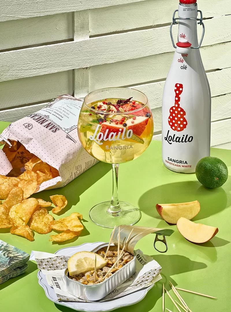 Verre de sangria blanche Lolailo avec bouteille, chips gourmet et fruits frais, idéal pour un moment d'été.