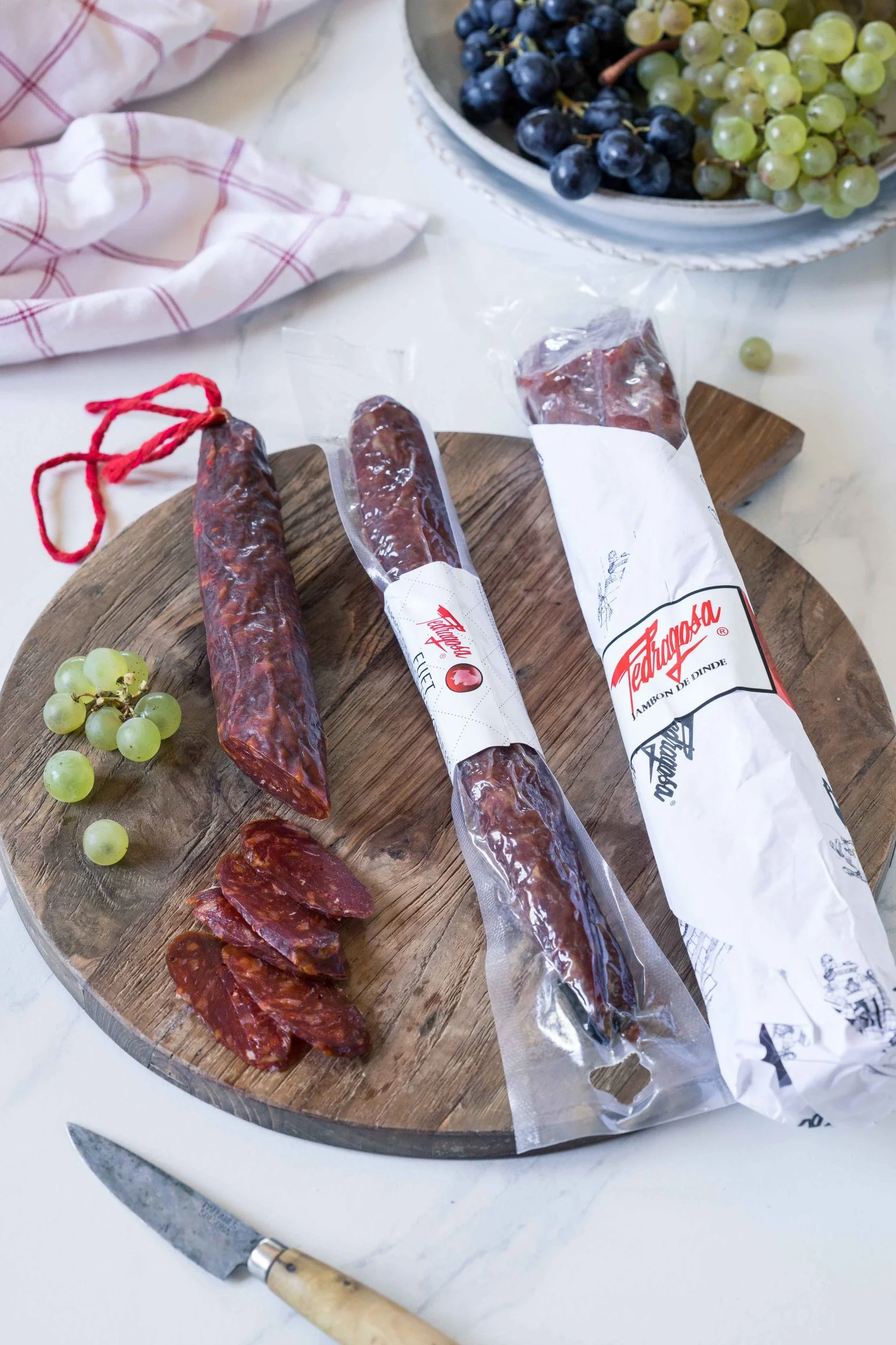 Sélection de fuet et saucisson catalans sur planche en bois, accompagnés de raisins frais.