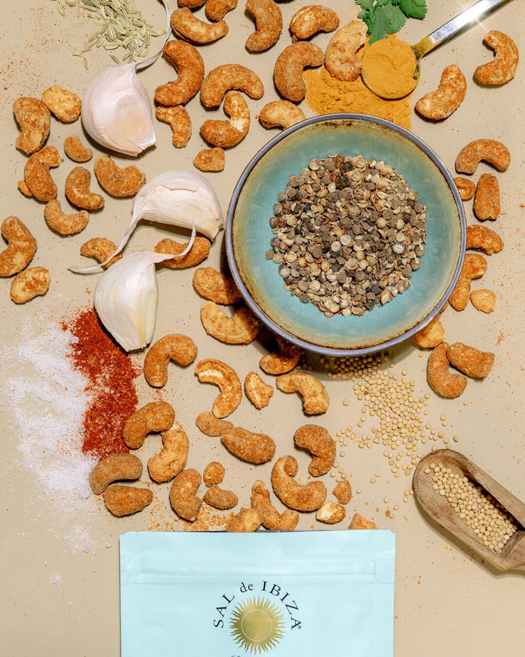Noix de cajou créoles Sal de Ibiza présentées avec leurs épices d’assaisonnement : ail, graines de moutarde, poivre, piment, fenouil et fleur de sel, autour d’un sachet turquoise de la marque.