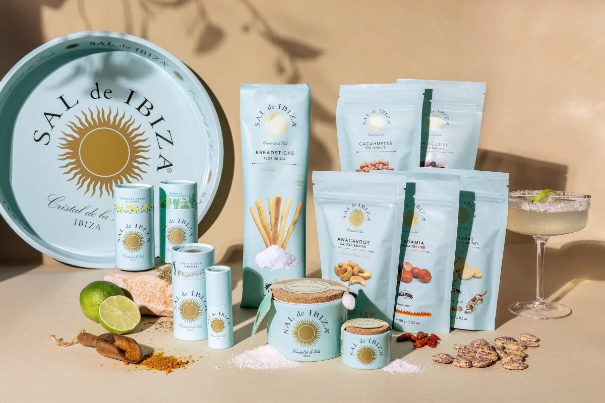 Gamme complète Sal de Ibiza : fleur de sel en pot céramique, sels aromatisés en tubes, gressins artisanaux et fruits à coque apéritifs, sur fond ensoleillé avec verre cocktail salé.