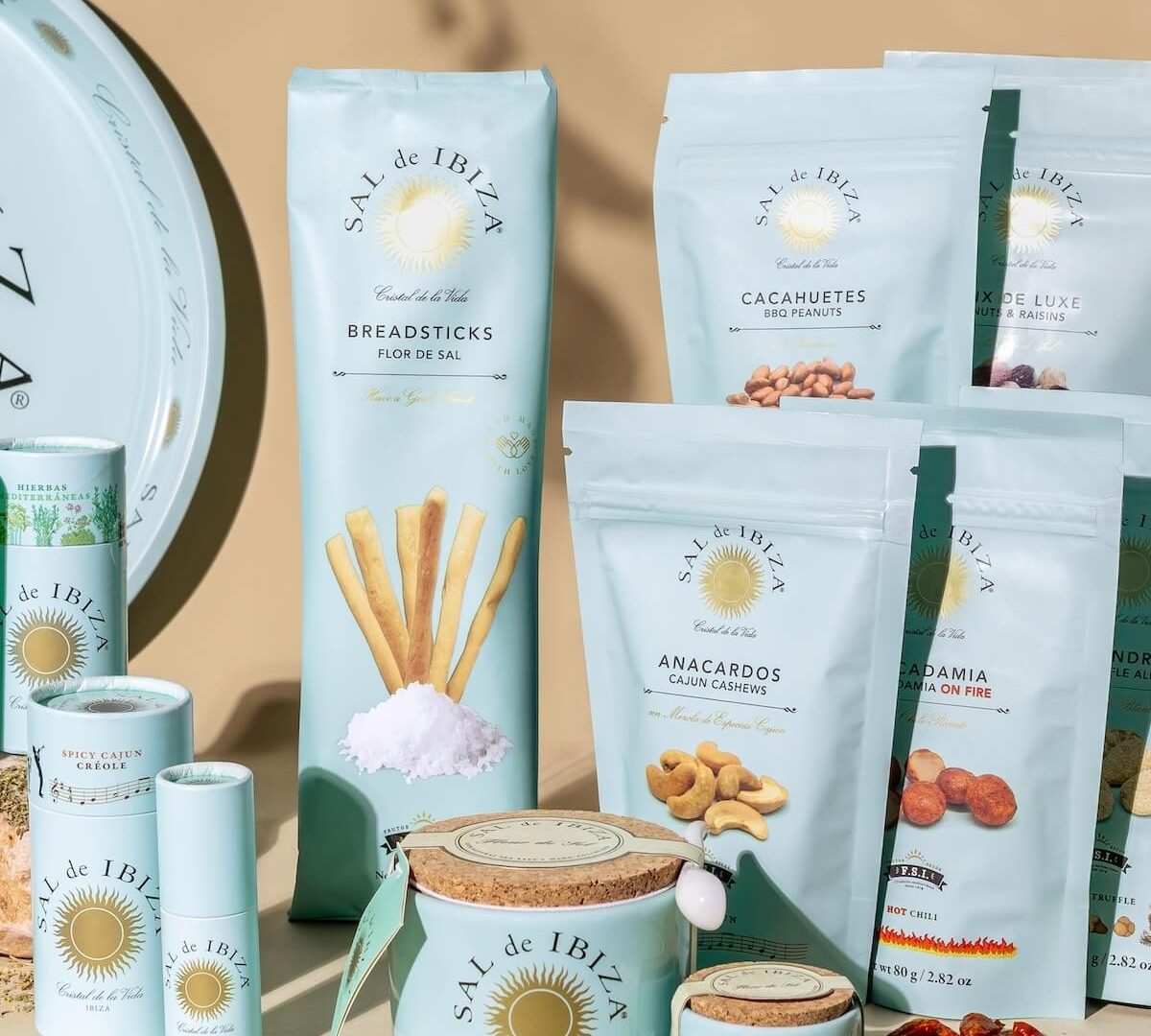 Sachets de fruits secs Sal de Ibiza : amandes, noix de cajou, cacahuètes et macadamia aux épices, avec pots de fleur de sel en céramique et cocktail en arrière-plan.