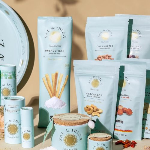 Sachets de fruits secs Sal de Ibiza : amandes, noix de cajou, cacahuètes et macadamia aux épices, avec pots de fleur de sel en céramique et cocktail en arrière-plan.