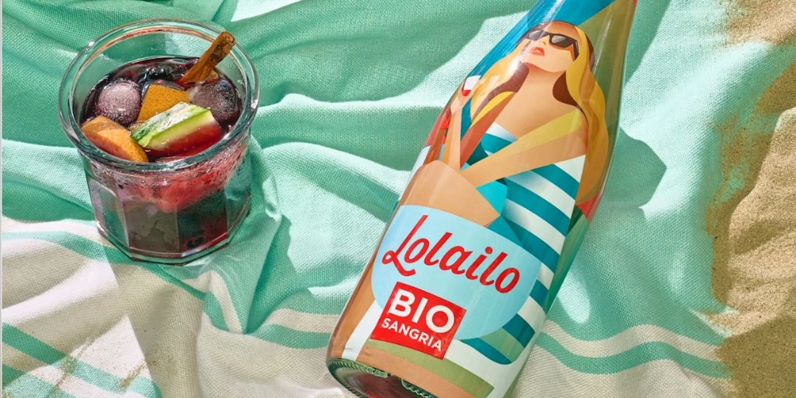 Bouteille de sangria bio Lolailo posée sur une serviette de plage avec un verre de sangria rouge