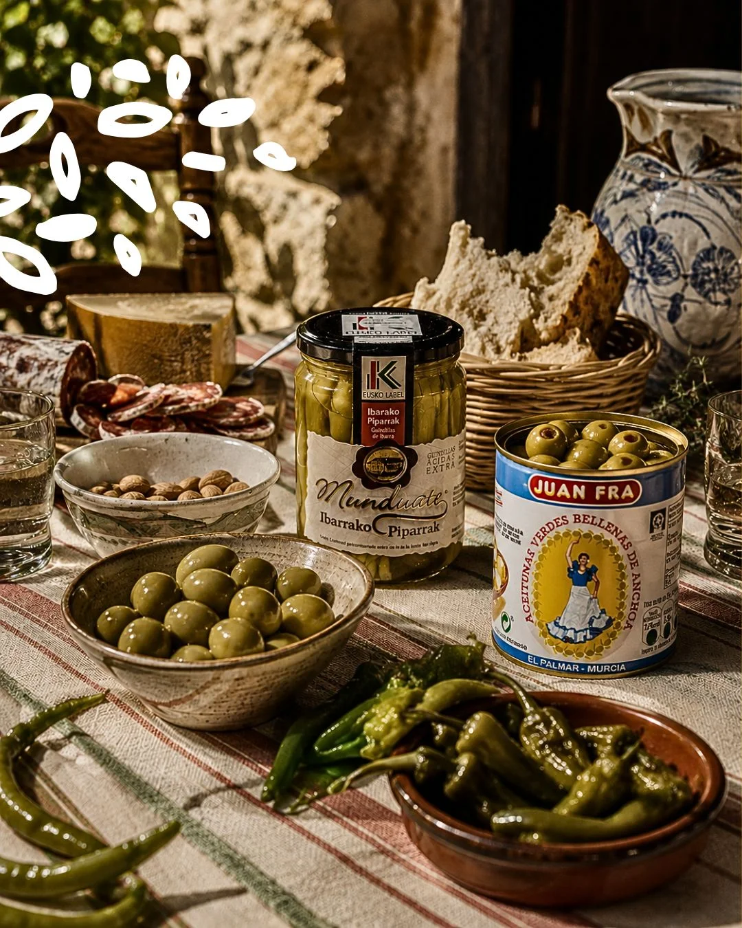 Table rustique dressée avec piments d'Ibarra, olives, charcuterie, fromages, pain et huile d'olive dans une ambiance basque.