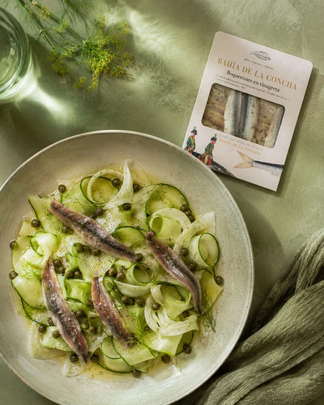 Vue de dessus d'une salade de fenouil, courgette en rubans et câpres surmontée de filets de boquerones, avec la boîte Bahía de la Concha Boquerones en vinagreta posée à côté sur un fond vert sauge