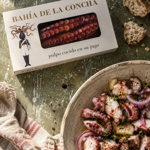 Étui de poulpe précuit Bahía de la Concha posé sur une table en bois patiné vert, à côté d'un pain de campagne et d'un torchon en lin rayé rouge, avec en amorce une salade de poulpe et pommes de terre
