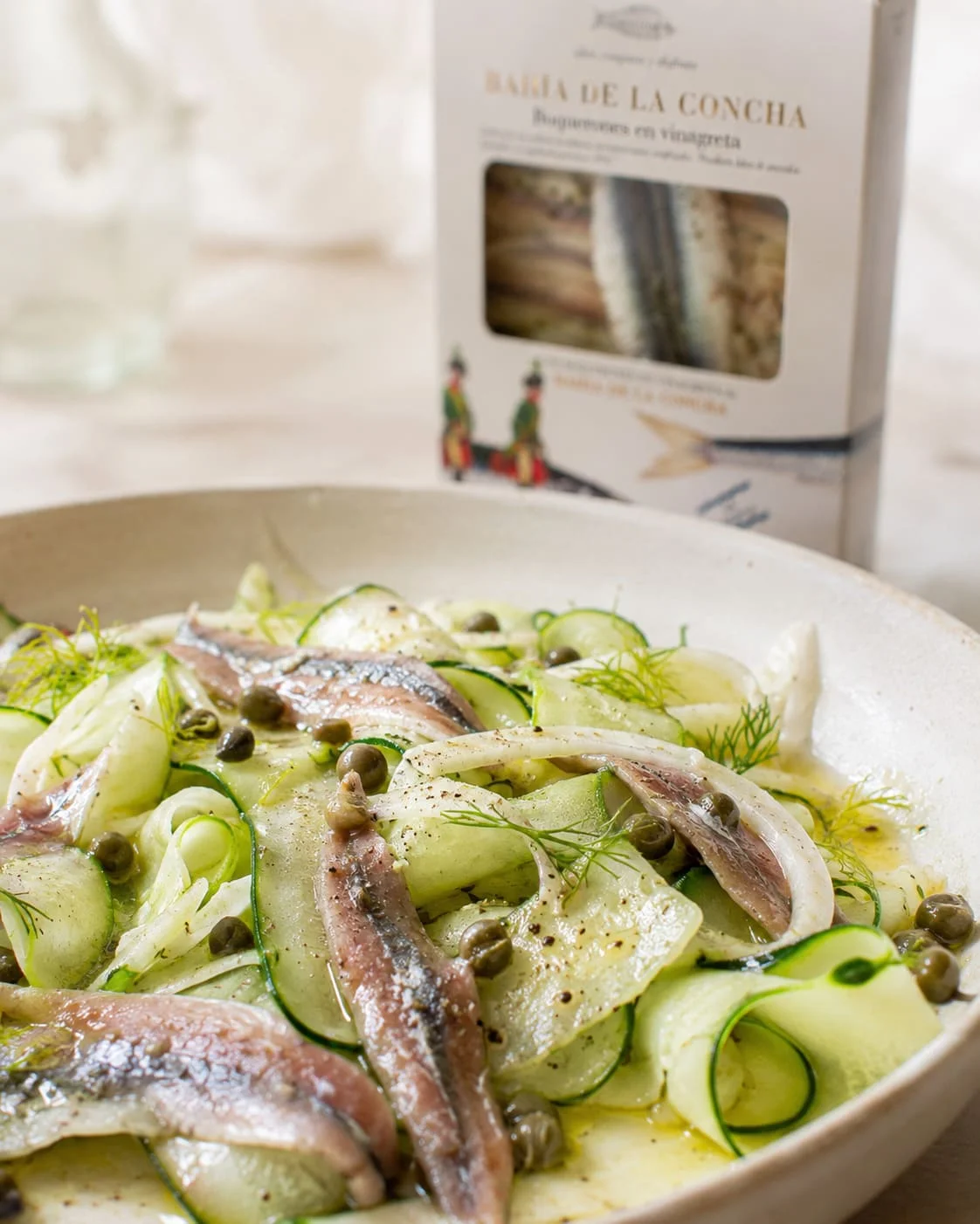 Gros plan sur une assiette de boquerones, fenouil et courgette en rubans, avec la boîte Bahía de la Concha floutée en arrière-plan et un brin d'aneth