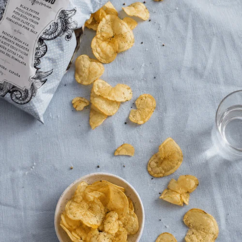 Sachet de chips San Nicasio ouvert, chips renversées sur un linge bleu clair à côté d'un bol en céramique et d'un verre d'eau