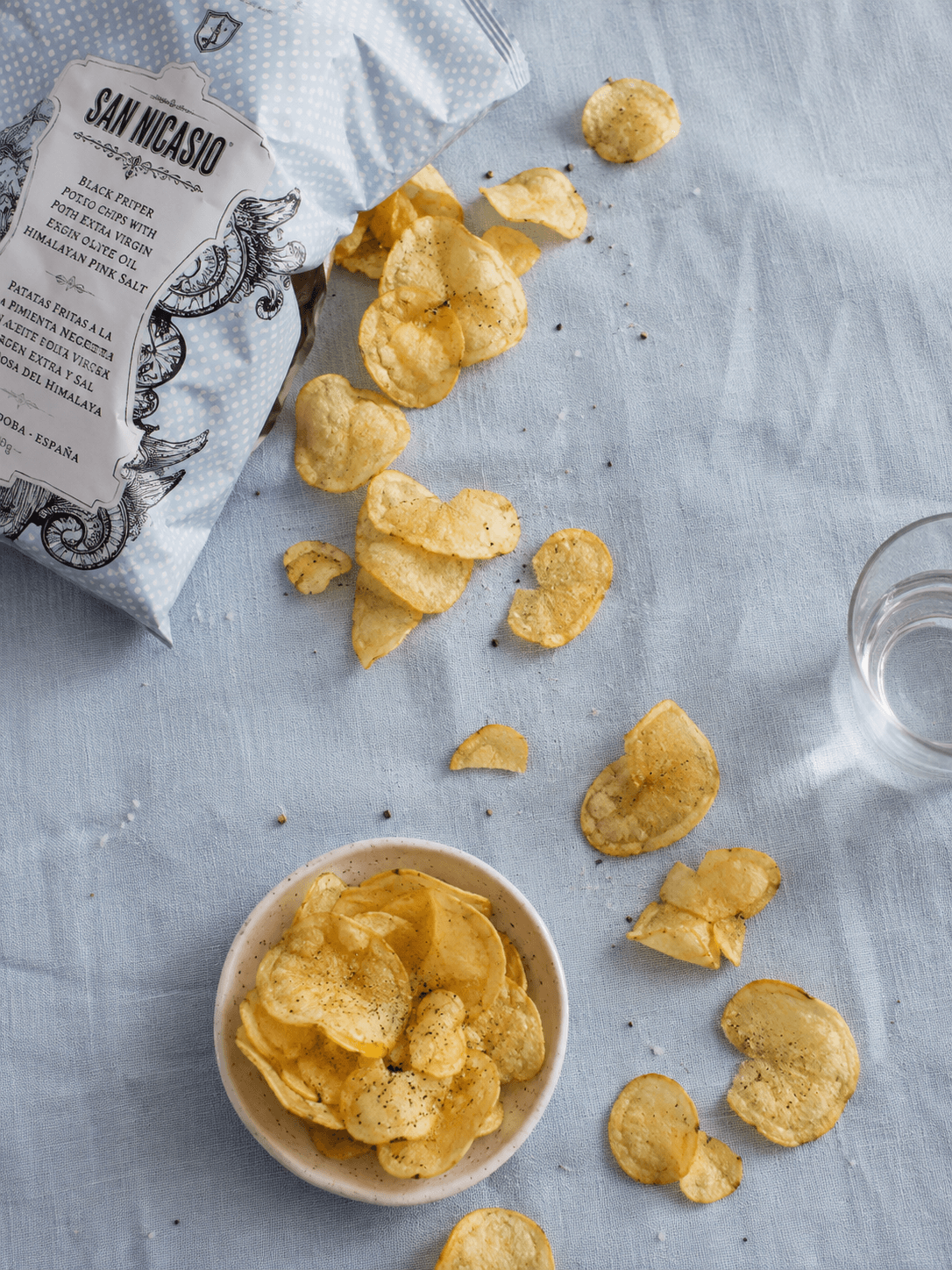Sachet de chips San Nicasio ouvert, chips renversées sur un linge bleu clair à côté d'un bol en céramique et d'un verre d'eau