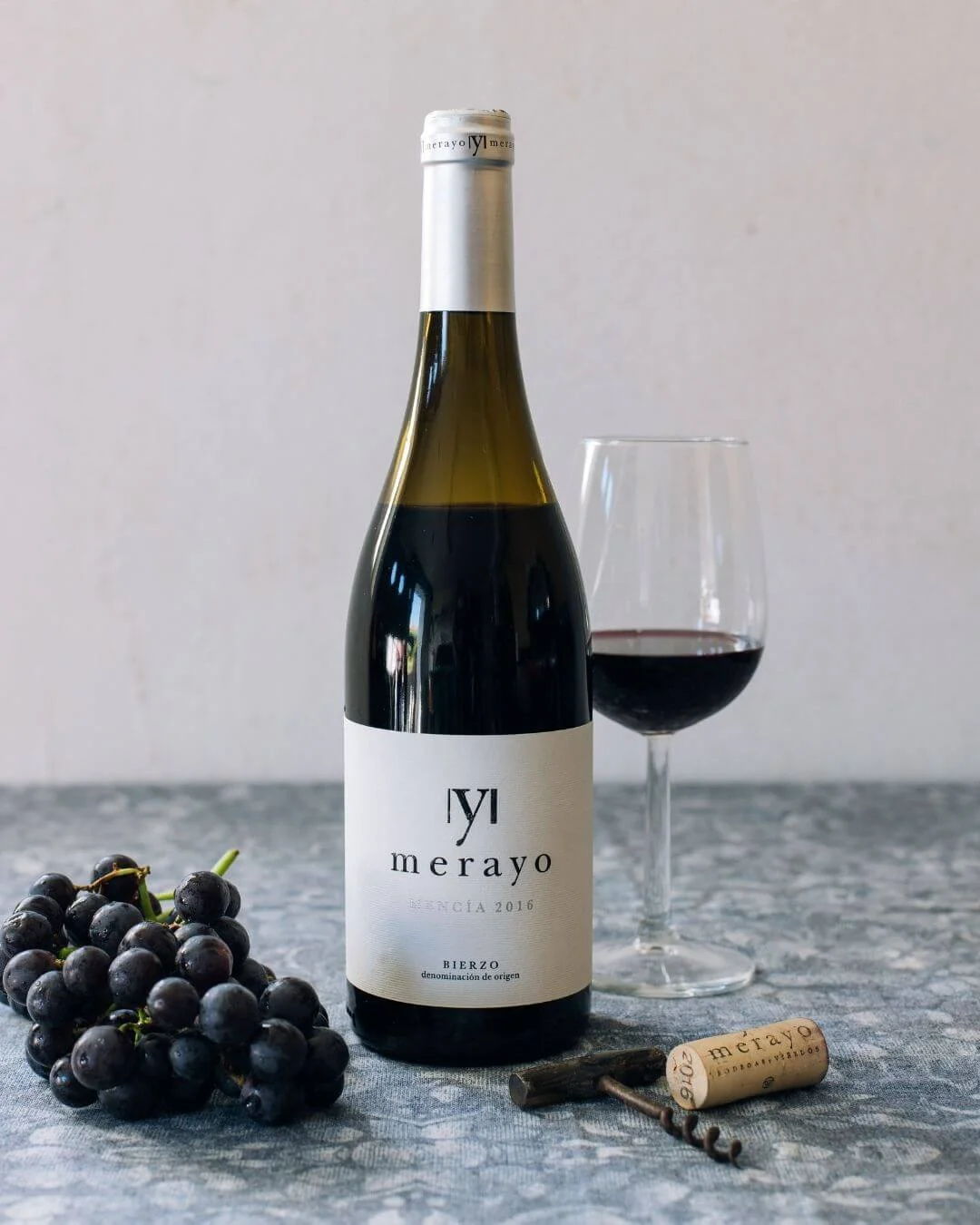 Bouteille de vin rouge Mencía Bodegas Merayo, AOP Bierzo, servie dans un verre à vin, avec une grappe de raisin noir et un tire-bouchon sur une nappe à motifs