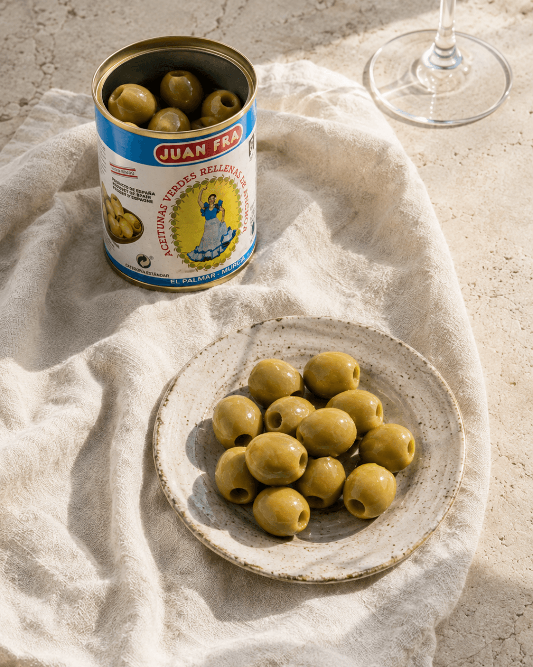 Boîte ouverte d'olives Manzanilla Juan Fra farcies à l'anchois, servies sur une assiette en céramique avec un verre de vin