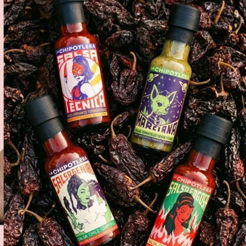 Quatre petites bouteilles de sauce piquante La Chipotlera aux étiquettes illustrées (Técnica, Marciana, Genial, Fogosa) posées sur un lit de piments chipotle séchés