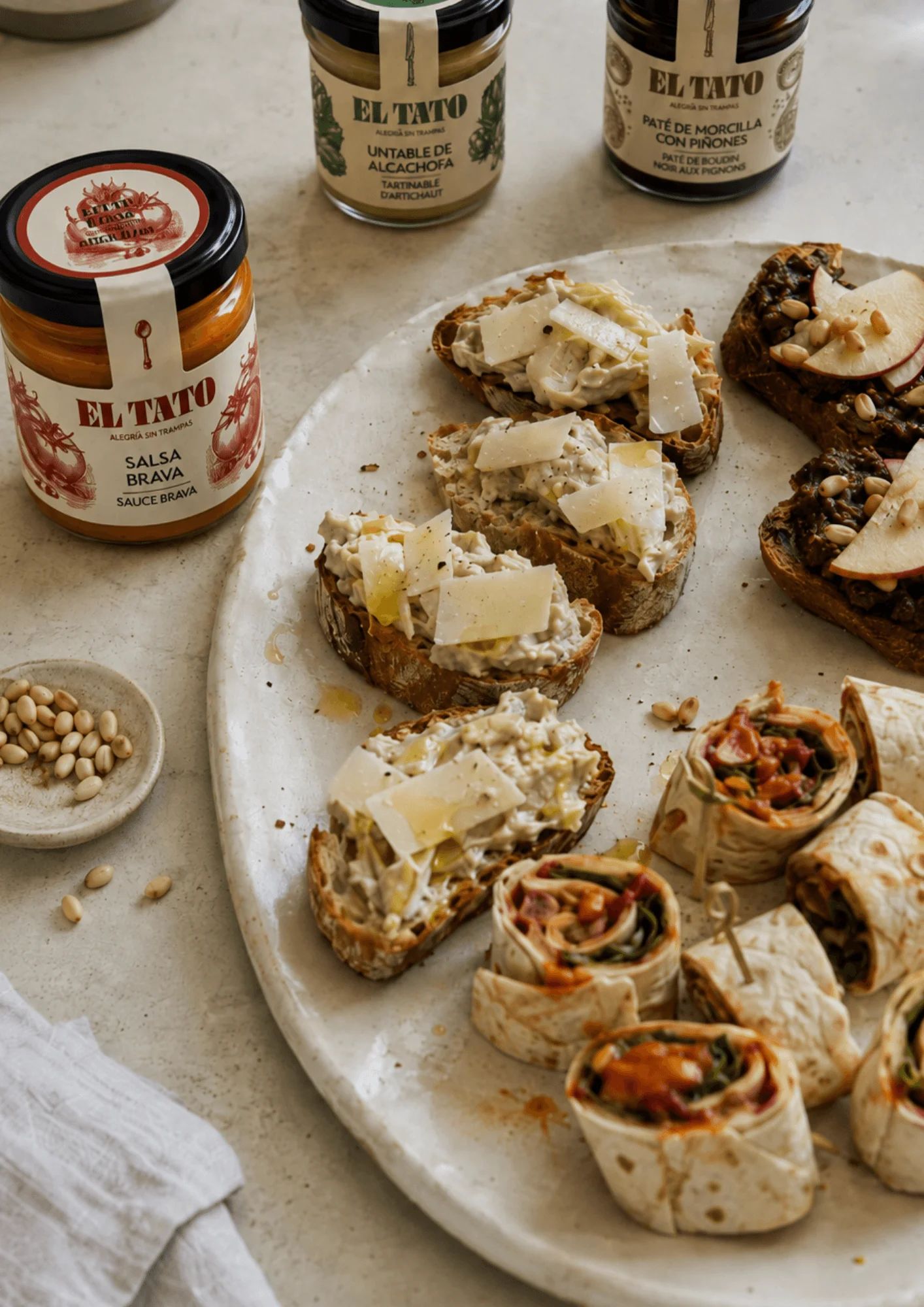 Assiette de toasts garnis de tartinable d'artichaut et copeaux de parmesan, et de toasts garnis de pâté de morcilla aux pignons et pomme, avec en arrière-plan les bocaux El Tato correspondants et la salsa brava