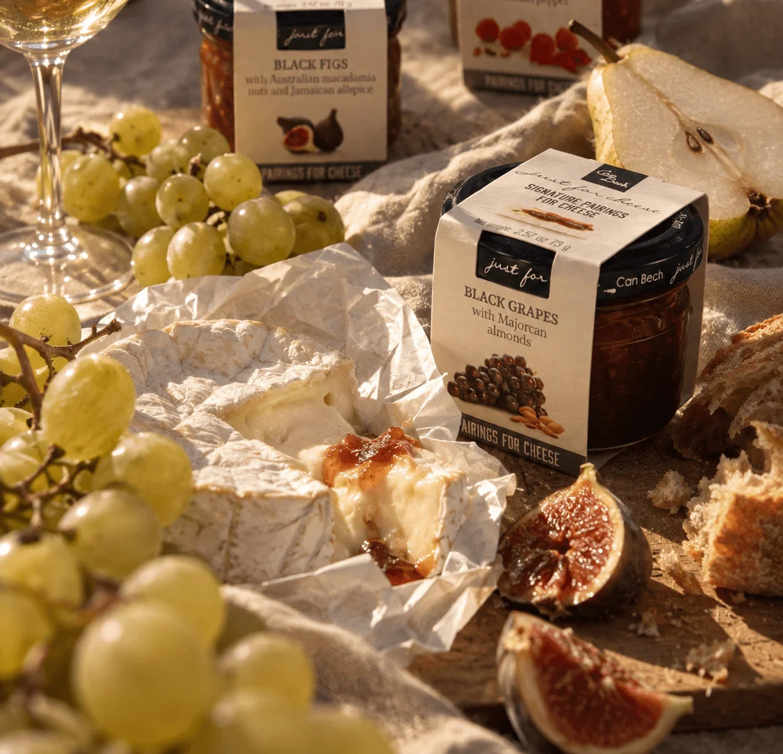 Plateau avec un camembert ouvert nappé de confiture de figues Can Bech, entouré de grappes de raisin blanc, de figues fraîches coupées, de pain de campagne et d'un verre de vin blanc, avec les pots Black Figs, Raspberries et Black Grapes de la gamme Just for Cheese en arrière-plan.