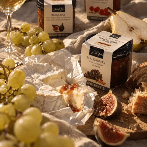 Plateau avec un camembert ouvert nappé de confiture de figues Can Bech, entouré de grappes de raisin blanc, de figues fraîches coupées, de pain de campagne et d'un verre de vin blanc, avec les pots Black Figs, Raspberries et Black Grapes de la gamme Just for Cheese en arrière-plan.