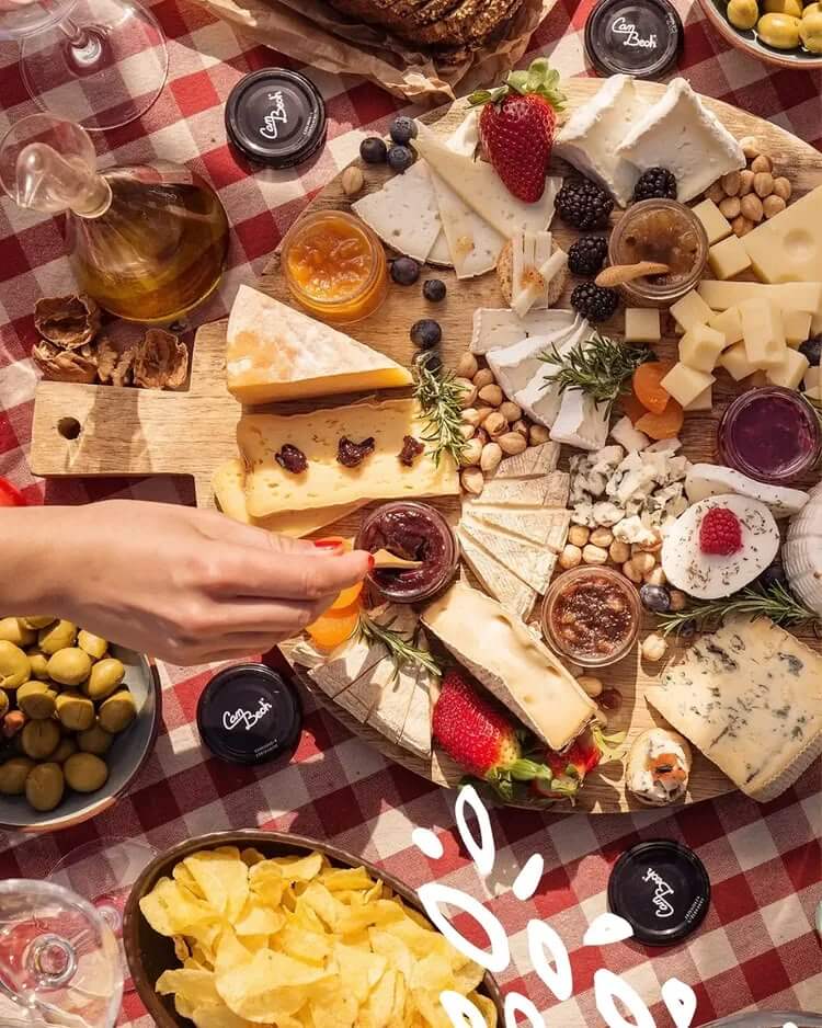 Vue du dessus d'un plateau de fromages sur nappe à carreaux rouge et blanc, avec plusieurs types de fromages, fruits rouges, noix, plusieurs pots Can Bech ouverts, une main qui prélève une spécialité à la cuillère, accompagnés d'olives, de chips et d'un carafon d'huile.