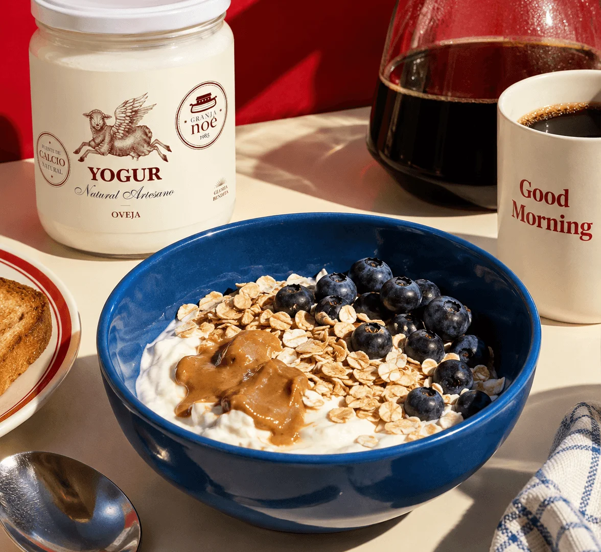 Bol bleu de yaourt de brebis garni de flocons d'avoine, de myrtilles et de beurre de cacahuète, à côté d'un pot de yaourt Granja Noé, d'une cafetière et d'une tasse