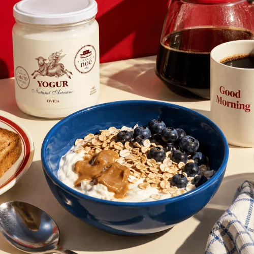 Bol bleu de yaourt de brebis garni de flocons d'avoine, de myrtilles et de beurre de cacahuète, à côté d'un pot de yaourt Granja Noé, d'une cafetière et d'une tasse