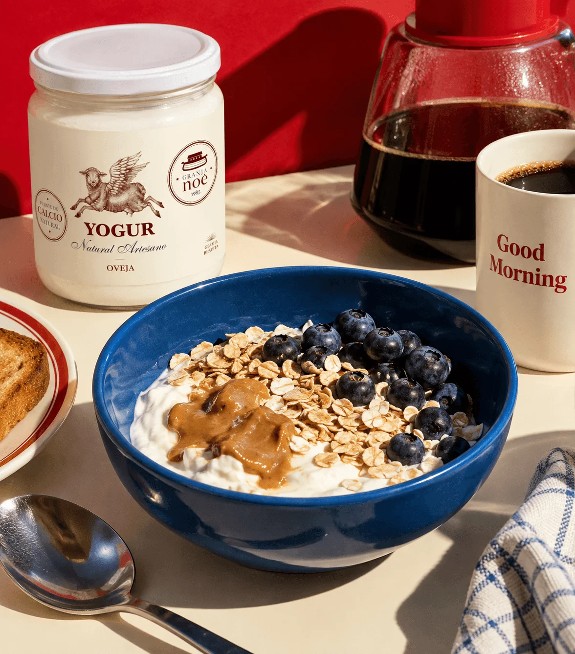 Bol bleu de yaourt de brebis garni de flocons d'avoine, de myrtilles et de beurre de cacahuète, à côté d'un pot de yaourt Granja Noé, d'une cafetière et d'une tasse