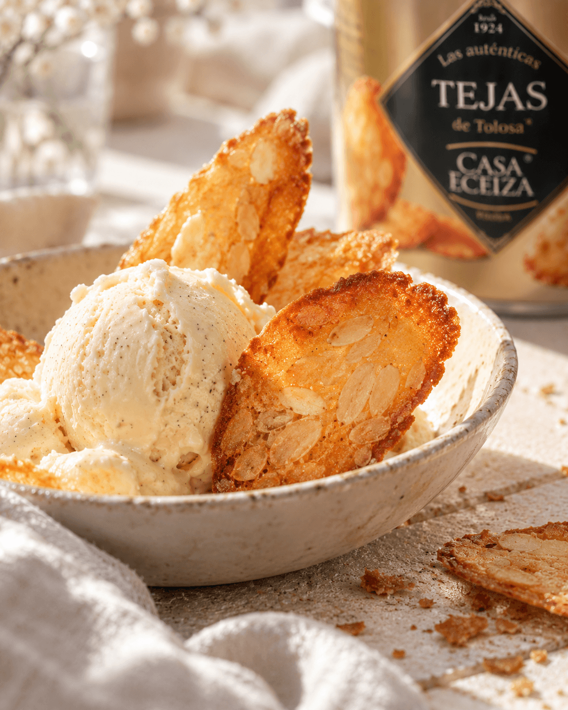 Boule de glace vanille piquée de tuiles aux amandes dans un bol en grès, avec la boîte « Las auténticas Tejas de Tolosa » Casa Eceiza, depuis 1924, en arrière-plan