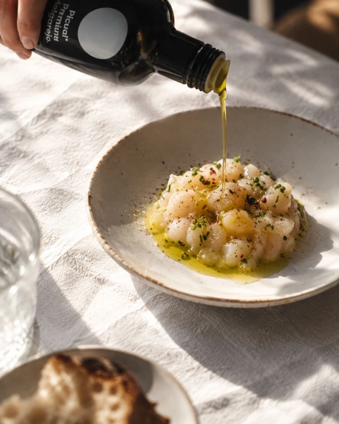 Filet d'huile d'olive vierge extra Melgarejo Picual Premium versé sur un tartare de la mer, avec pain et verre d'eau sur une nappe en lin