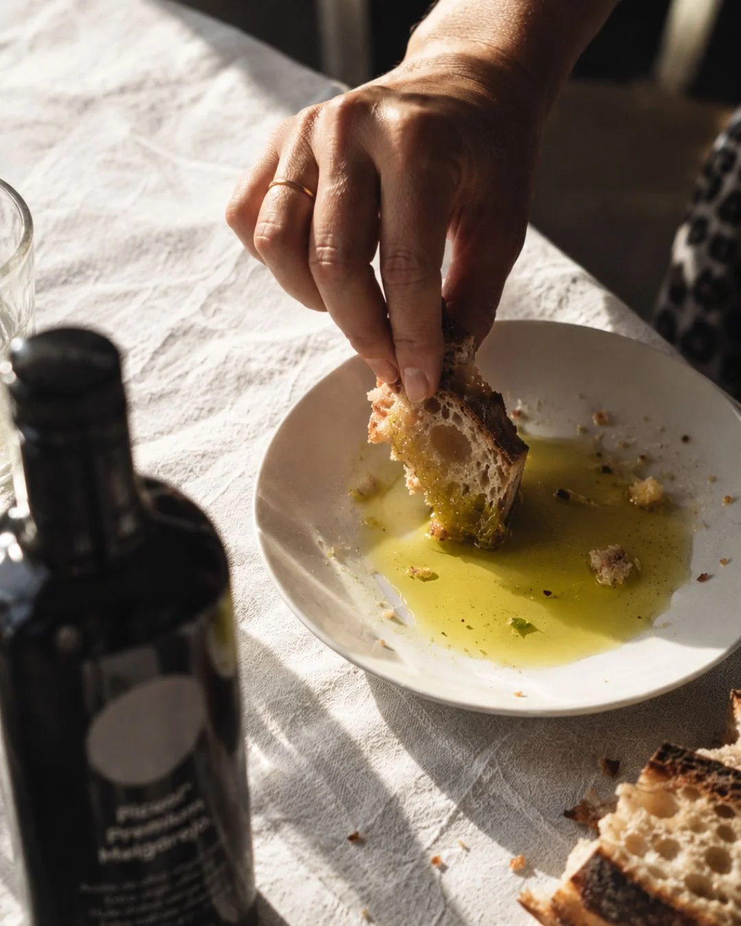 Main trempant un morceau de pain de campagne dans une assiette d'huile d'olive Picual Melgarejo, bouteille au premier plan