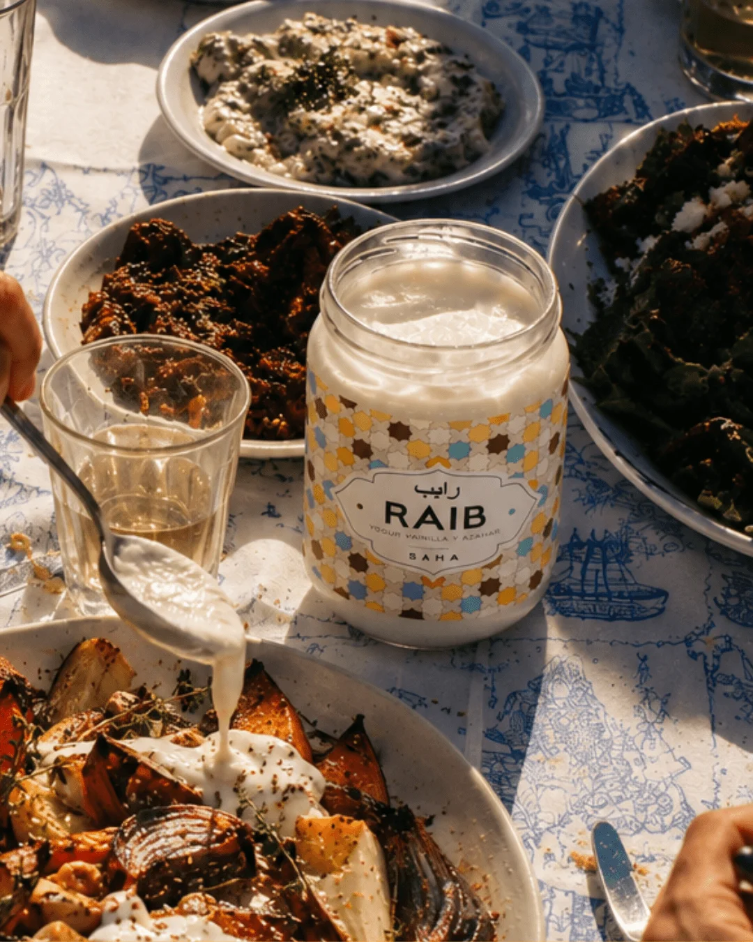 Pot ouvert de raïb sur une table de partage, avec une cuillère de laitage fermenté versée sur un plat de légumes rôtis