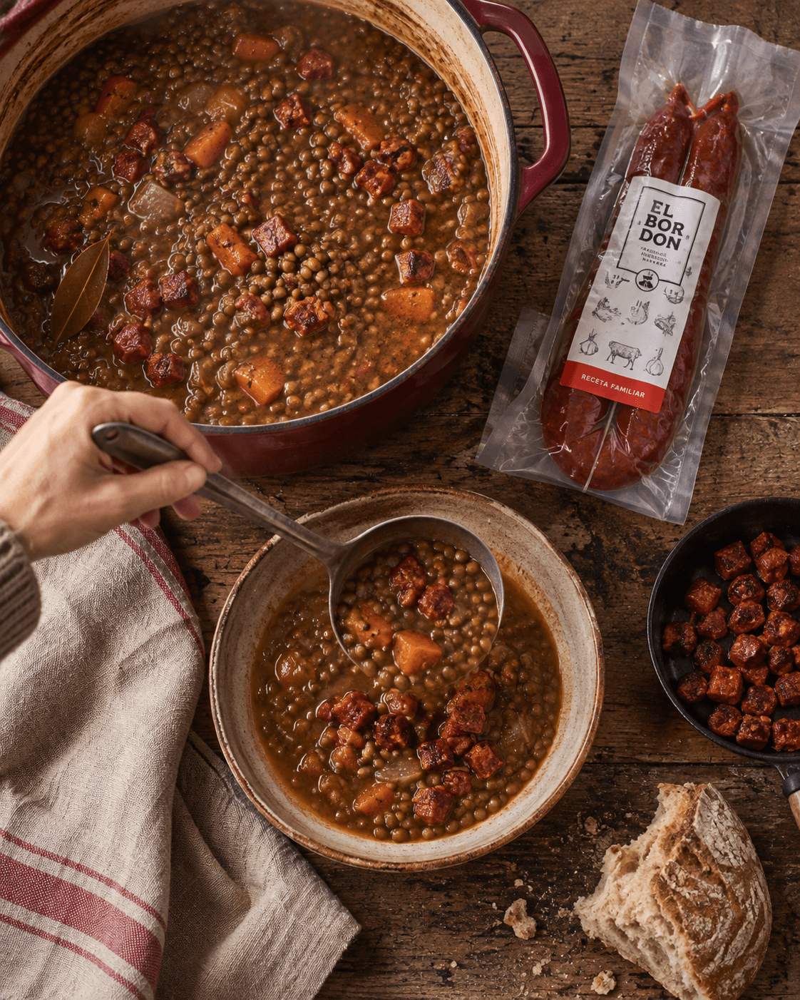 Soupe de lentilles au chorizo servie à la louche dans un bol, avec une cocotte rouge, un chorizo El Bordón sous vide et des dés de chorizo poêlés