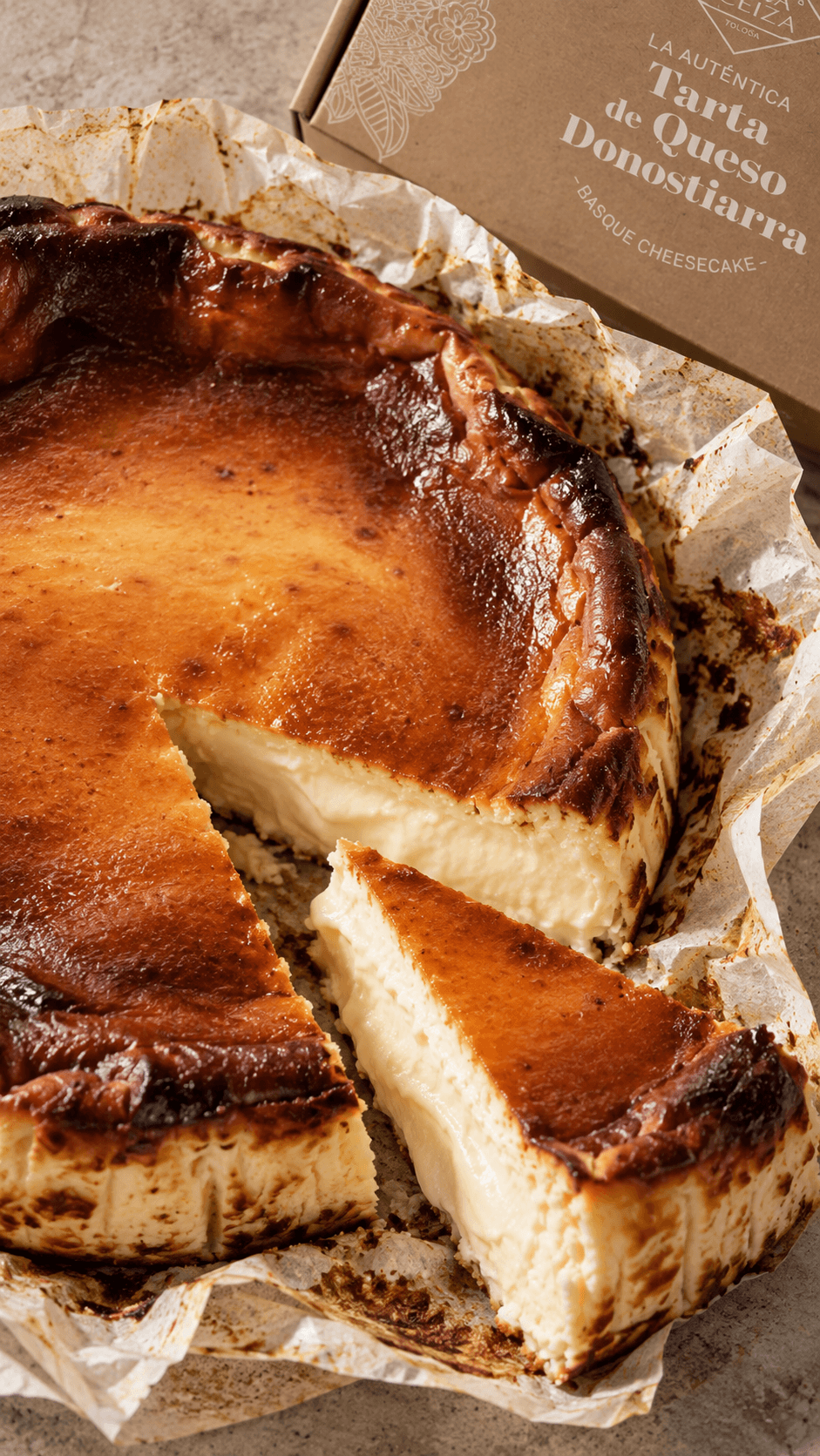 Cheesecake basque à la surface caramélisée sur papier sulfurisé, une part découpée laissant voir le cœur crémeux, et la boîte kraft « La auténtica Tarta de Queso Donostiarra »